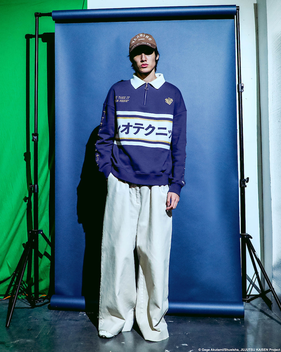 Male modeling Atsuko Kento Nanami Blue & Yellow Quarter-Zip Pullover with a navy blue base, white and yellow stripes, Japanese text across the chest, and a white collar with a ring zipper on a blue, and green background.