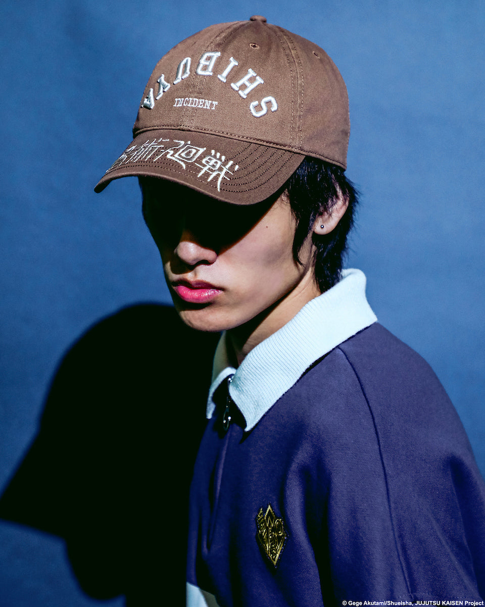 Male modeling brown Shibuya Incident Hat inspired by Jujutsu Kaisen, featuring white embroidered text and Japanese characters on the front and brim in front of a blue background.