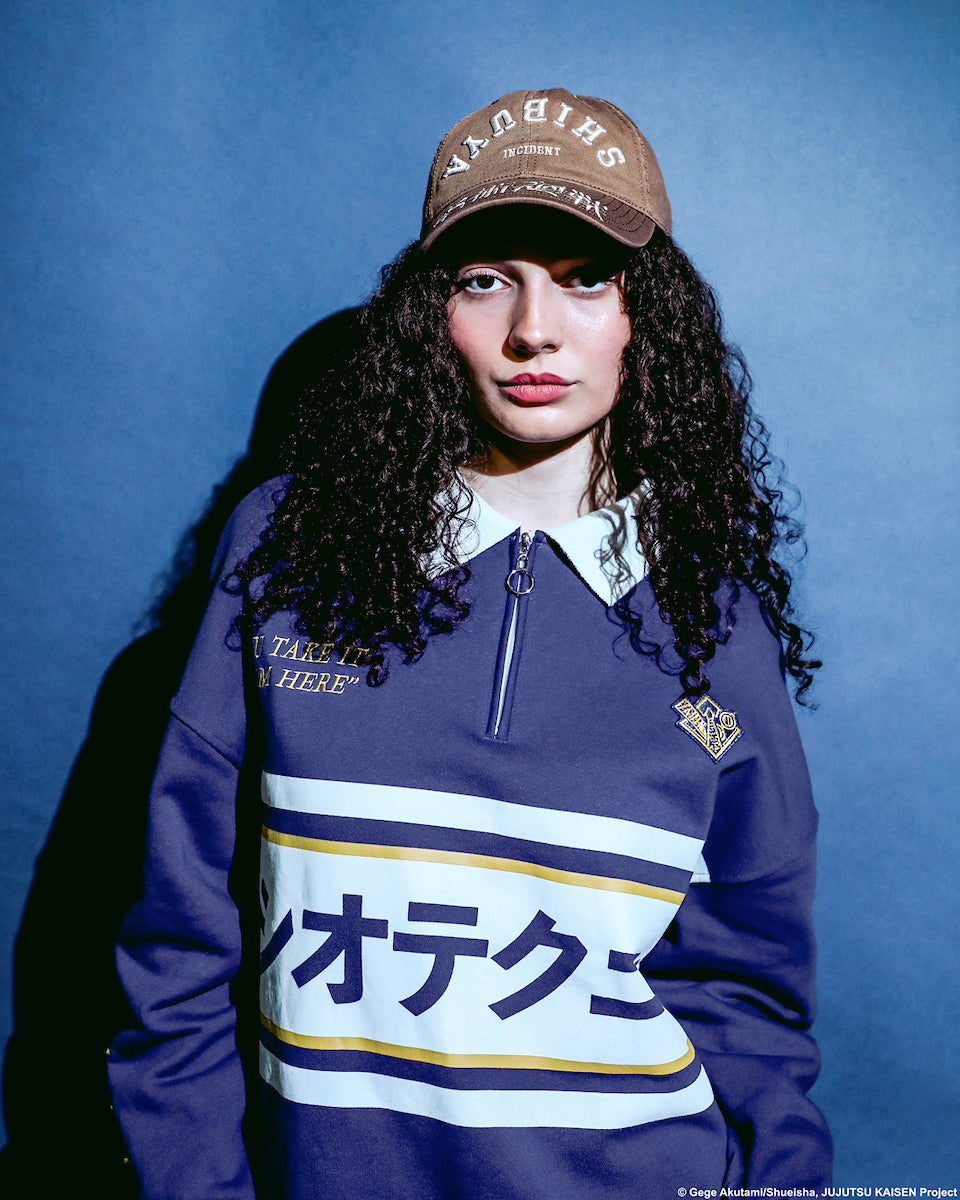 Close-up of a female modeling Atsuko Kento Nanami Blue & Yellow Quarter-Zip Pullover with a navy blue base, white and yellow stripes, Japanese text across the chest, and a white collar with a ring zipper on a pale blue background.