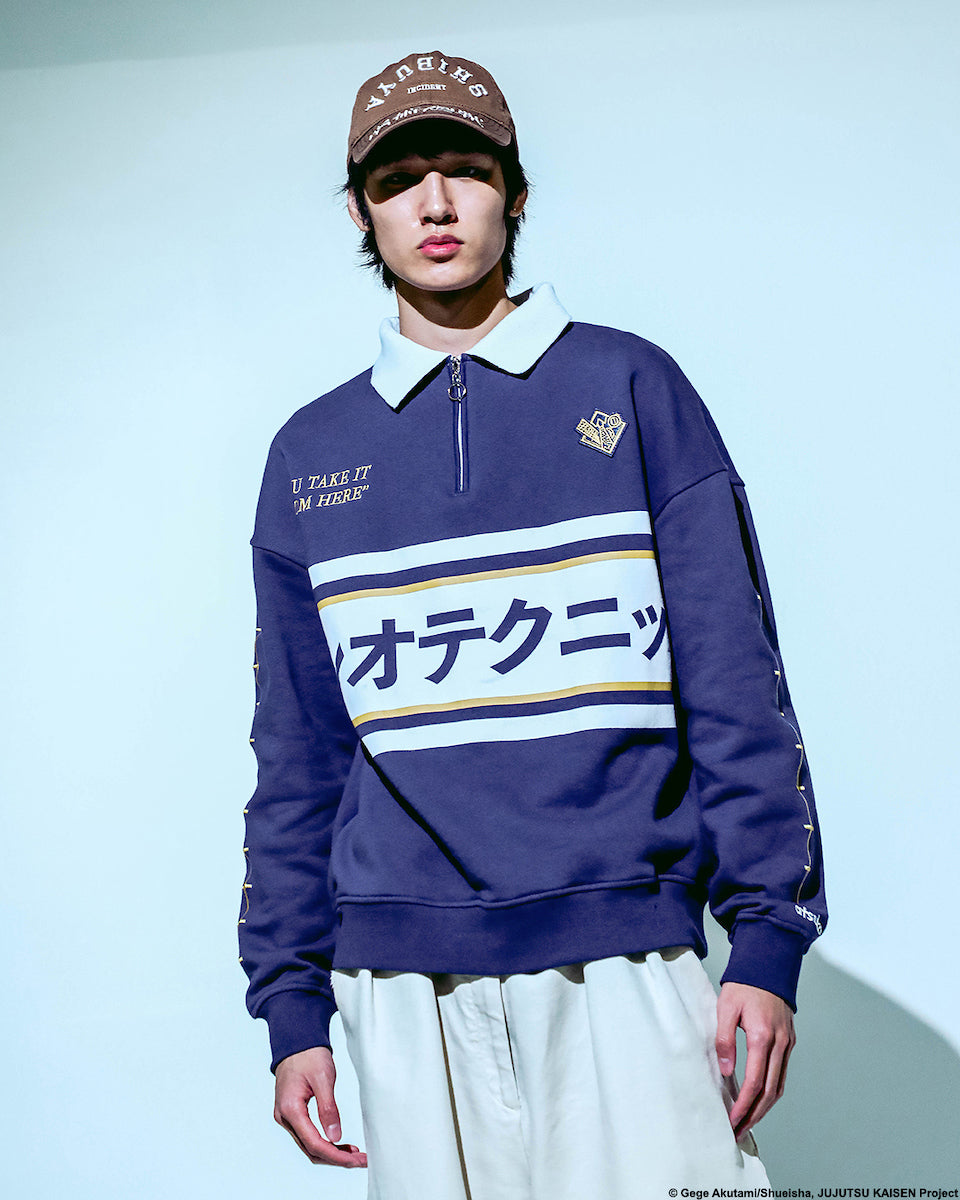 Male modeling Atsuko Kento Nanami Blue & Yellow Quarter-Zip Pullover with a navy blue base, white and yellow stripes, Japanese text across the chest, and a white collar with a ring zipper on a pale blue background.